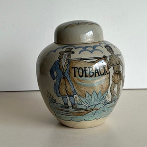 Pode incluir: Um pote de cerâmica com tampa, pintado com uma cena de duas figuras segurando uma faixa que diz "TOEBAC". O pote é decorado com detalhes azuis e marrons.