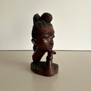 Peut inclure: Une sculpture en bois brun foncé représentant la tête et les épaules d'une personne. La personne a les cheveux tressés et regarde sur le côté. La sculpture est sculptée dans un style réaliste.