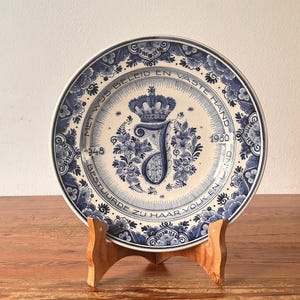 May include: A blue and white decorative plate with floral and crown designs, displayed on a wooden stand. The plate features text and the year 1948 and 1980. The plate has a silver rim.