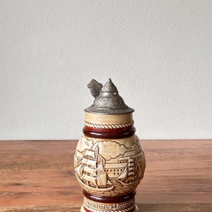 Könnte beinhalten: Ein Keramik-Bierkrug mit einem silbernen Deckel und einer detaillierten Szene eines Schiffes, das in einen Hafen segelt. Der Krug hat ein braunes und rotes Seil-Design um die Mitte und die Worte "Barkentine J. Briggand" am Boden.