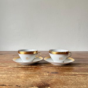 Puede incluir: Dos tazas de té y platillos blancos con adornos y detalles dorados. Las tazas tienen una banda dorada alrededor del borde y un asa dorada. Los platillos tienen un borde dorado. Sobre una superficie de madera.