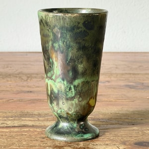 Puede incluir: Una copa de cerámica con un esmalte moteado en tonos verde bosque, marrón oscuro y toques de amarillo. El recipiente tiene un cuerpo cilíndrico cónico que se ensancha ligeramente en el borde y descansa sobre una base circular de pedestal.