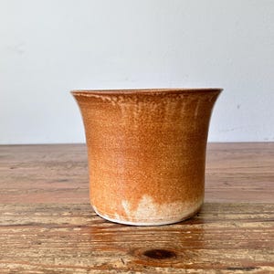 Pode incluir: Um vaso de cerâmica castanho artesanal com uma borda alargada. O vaso tem uma textura salpicada e uma base mais clara, sem vidrado. Assenta numa superfície de madeira, mostrando os seus tons terrosos e o seu design simples.