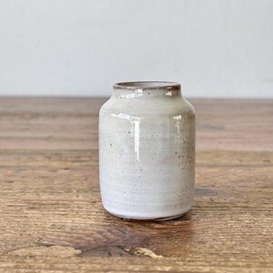 Pode incluir: Um pequeno vaso de cerâmica branco-sujo com acabamento salpicado. O vaso tem um corpo cilíndrico com uma borda ligeiramente alargada e uma borda marrom escura. O vaso é exibido em uma superfície de madeira.