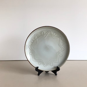 Könnte beinhalten: Ein weißer Keramikteller mit braunem Rand und einem sternförmigen Design. Der Teller steht auf einem schwarzen Ständer.