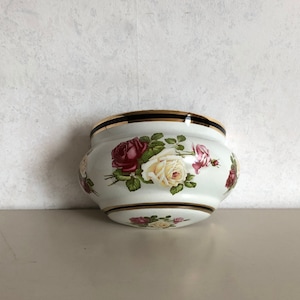 Könnte beinhalten: Weißer Keramik-Pflanztopf mit goldfarbenem Rand und schwarzem Streifen. Der Topf hat ein Blumenmuster mit rosa, weißen und gelben Rosen.