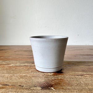 May include: A small, light gray ceramic cup with a slightly flared shape. The cup has a simple, minimalist design and sits on a wooden surface. The cup is approximately 3 inches tall and 3 inches wide.