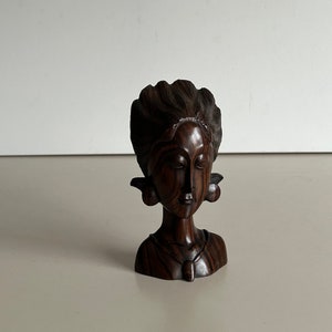 Op de afbeelding: Een donkerbruine houten sculptuur van het hoofd en de schouders van een vrouw. De sculptuur heeft gedetailleerde gravures van het haar, gezicht en oorbellen. Een ketting met een hanger is ook zichtbaar. Het stuk staat tegen een neutrale achtergrond.