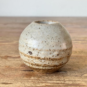 May include: A small, spherical ceramic vase with a speckled, light grey exterior and a brown, banded base. The vase has a small opening at the top and sits on a wooden surface. The pottery has a rustic, handmade appearance.