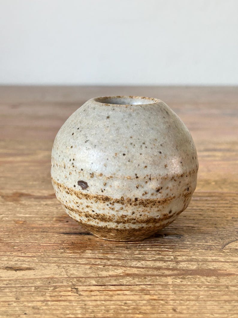 May include: A small, spherical ceramic vase with a speckled, light grey exterior and a brown, banded base. The vase has a small opening at the top and sits on a wooden surface. The pottery has a rustic, handmade appearance.