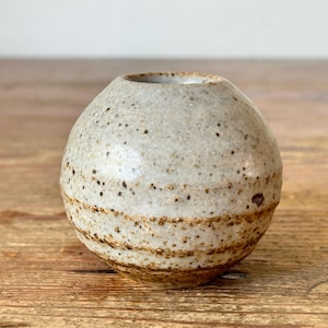 May include: A small, spherical ceramic vase with a speckled, off-white glaze. The vase has a narrow opening and a textured surface with brown bands near the base. The handmade pottery is suitable for displaying small flowers or as a decorative accent.
