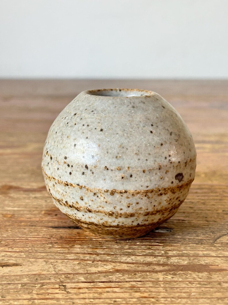 May include: A small, spherical ceramic vase with a speckled, off-white glaze. The vase has a narrow opening and a textured surface with brown bands near the base. The handmade pottery is suitable for displaying small flowers or as a decorative accent.