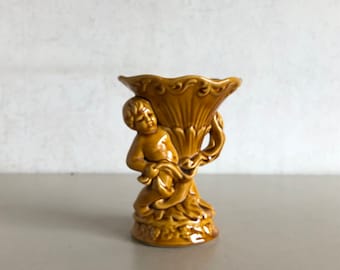 Może przedstawiać: Brązowy ceramiczny wazon z figurką cheruba trzymającego kwiatowy wzór. Wazon ma falisty brzeg i ozdobną podstawę.