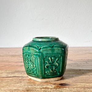 Pode incluir: Um pequeno recipiente de cerâmica hexagonal com um esmalte verde esmeralda profundo. O exterior apresenta painéis em relevo moldados que retratam motivos florais e botânicos. A base não é esmaltada, revelando uma textura de argila bege clara.