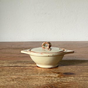 Peut inclure: Un petit bol en céramique rond avec un couvercle assorti, présentant une glaçure blanc cassé pâle et des accents brun rougeâtre rustiques sur les bords et la petite poignée en boucle sur le dessus. Le bol possède deux petites anses pointues.