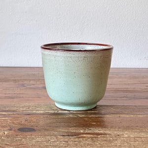 Può includere: Un vaso di ceramica verde chiaro con un bordo marrone. Il vaso ha una smaltatura macchiettata e una forma arrotondata.