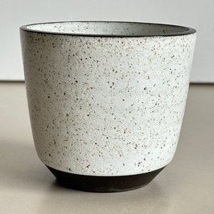 Puede incluir: Una taza de cerámica blanca moteada con base y borde marrón oscuro. La taza tiene una forma ligeramente cónica y una superficie texturizada, perfecta para café o té.