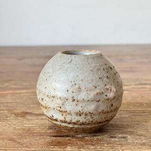 May include: A small, round, ceramic vase with a speckled, off-white exterior and a brown band near the base. The vase has a small opening at the top and sits on a wooden surface. The vase is approximately 7.6 cm in diameter.
