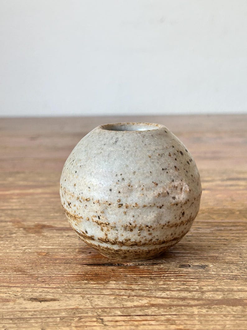May include: A small, round, ceramic vase with a speckled, off-white exterior and a brown band near the base. The vase has a small opening at the top and sits on a wooden surface. The vase is approximately 7.6 cm in diameter.