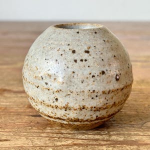 May include: A small, spherical ceramic vase with a speckled, off-white exterior and brown banded details. The vase has a small opening at the top. The pottery has a rustic, handmade appearance and sits on a wooden surface.