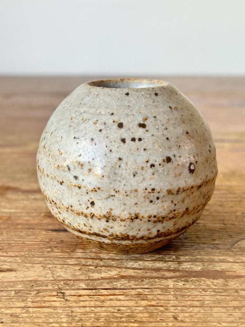 May include: A small, spherical ceramic vase with a speckled, off-white exterior and brown banded details. The vase has a small opening at the top. The pottery has a rustic, handmade appearance and sits on a wooden surface.