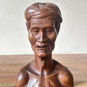 Op de afbeelding: Een houten buste van een man met een tulband. De sculptuur heeft een warme bruine kleur, met gedetailleerde gelaatstrekken en een getextureerde tulband. De buste staat op een houten oppervlak.