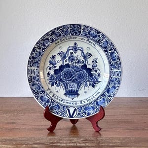May include: A blue and white Delftware plate with a floral design and a basket of flowers. The plate has the text "1945 27 October - 27 April 12% JJ Schiltmans - K.v. Rossum 1958" around the edge.