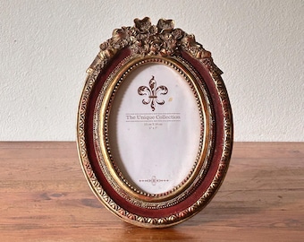 Könnte beinhalten: Ovaler Bilderrahmen mit bordeauxroter und goldener Oberfläche. Der Rahmen hat verzierte Details an der Oberseite und einen dekorativen Rand. Der Rahmen ist mit "The Unique Collection" beschriftet und misst 13 cm x 18 cm.