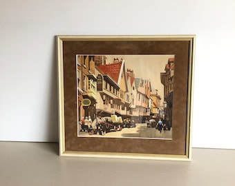 May include: A framed watercolor painting depicting a street scene in a European city. The painting features a row of old buildings with gabled roofs and a cobblestone street. There are horse-drawn carriages and people walking in the street.