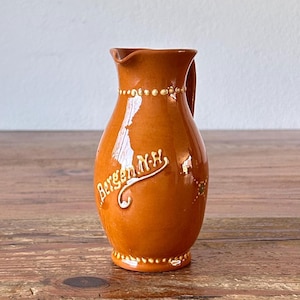 Puede incluir: Una jarra de cerámica marrón con asa y detalles blancos. La jarra tiene las palabras "Bergen, N.H." escritas en ella.