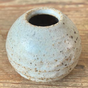 May include: A small, round ceramic vase with a light blue-grey speckled finish. The vase has a narrow opening and a dark interior. The surface has a textured appearance with small brown spots. Likely handmade.