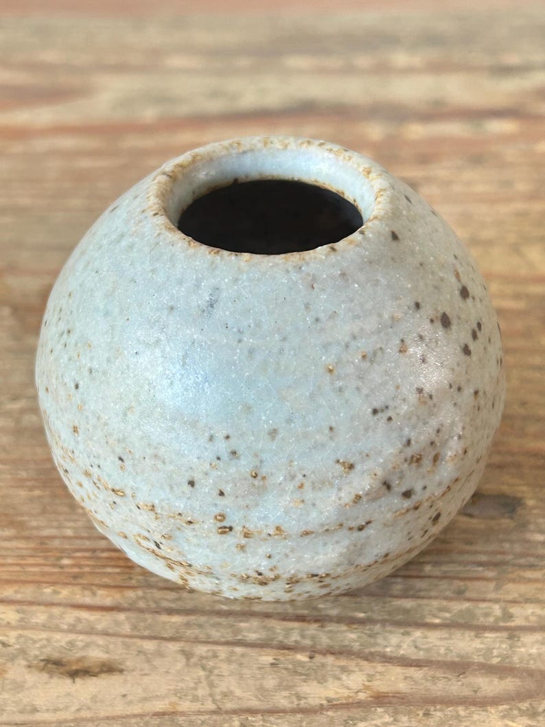 May include: A small, round ceramic vase with a light blue-grey speckled finish. The vase has a narrow opening and a dark interior. The surface has a textured appearance with small brown spots. Likely handmade.