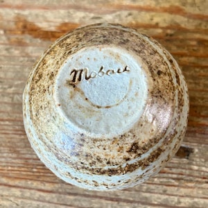 May include: A small, round ceramic bowl with a light blue interior and a textured, speckled exterior in shades of brown and tan. The word "Mobou" is inscribed on the base.