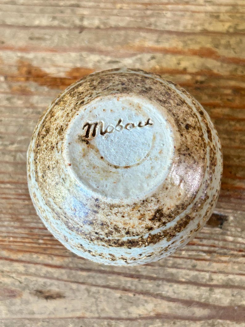 May include: A small, round ceramic bowl with a light blue interior and a textured, speckled exterior in shades of brown and tan. The word "Mobou" is inscribed on the base.