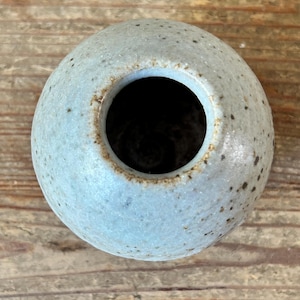 May include: A small, round, ceramic vase with a light blue glaze and speckled detailing. The vase has a wide opening and a dark interior. The handmade pottery is displayed on a wooden surface.