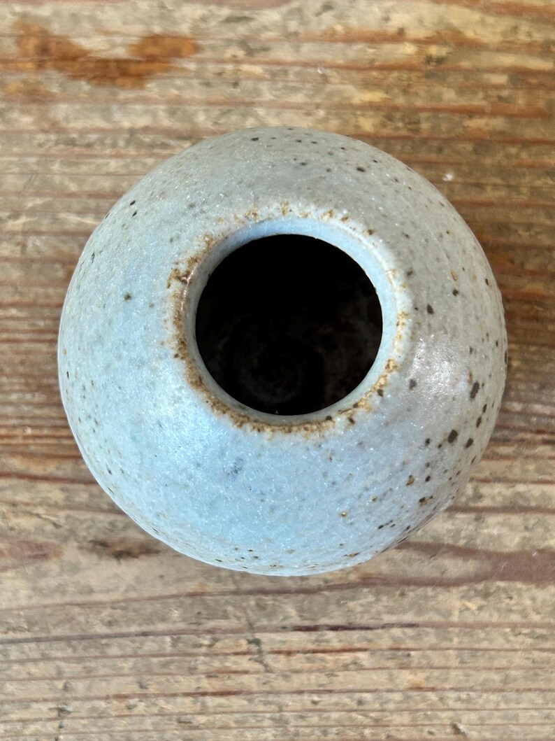 May include: A small, round, ceramic vase with a light blue glaze and speckled detailing. The vase has a wide opening and a dark interior. The handmade pottery is displayed on a wooden surface.