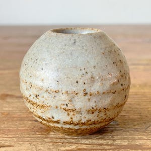 May include: A small, round ceramic vase with a speckled, off-white glaze and brown accents. The vase has a wide opening and sits on a wooden surface. The pottery has a rustic, handmade appearance.