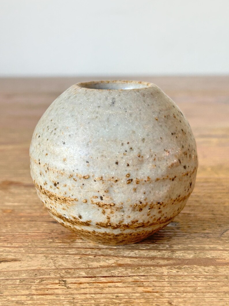 May include: A small, round ceramic vase with a speckled, off-white glaze and brown accents. The vase has a wide opening and sits on a wooden surface. The pottery has a rustic, handmade appearance.