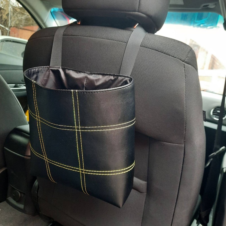 May include: Black leather car trash can with yellow stitching hanging from the headrest of a car seat.