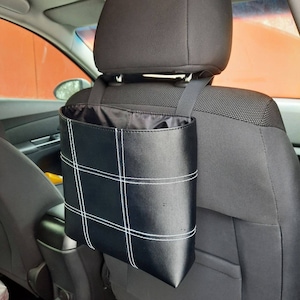 May include: Black leather car trash can with white stitching hanging from the headrest of a car seat.
