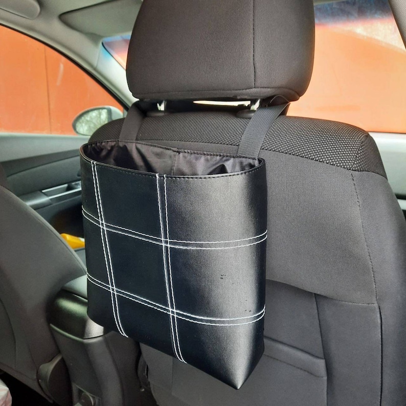May include: Black leather car trash can with white stitching hanging from the headrest of a car seat.