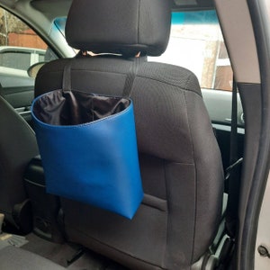 May include: A blue car trash can with black straps hangs from the headrest of a car seat. The trash can is made of a leather-like material and has a black lining.
