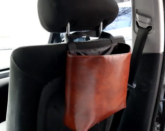 May include: A brown leather car trash can with a black fabric top hanging from the headrest of a car seat.
