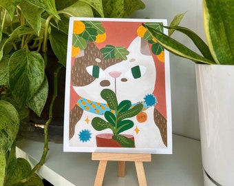May include: A whimsical art print of a cat with brown and white fur, wearing a blue collar with orange fruit. The cat is holding a potted green plant. The print is displayed on a small wooden easel, with green plants in the background.