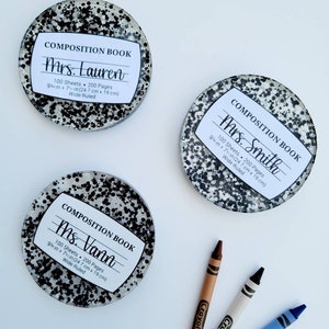 May include: Three round, black and white speckled coasters with white labels that say "Composition Book" and include the teacher's name, "Mrs. Lauren", "Mrs. Smith", and "Ms. Vann". Each label also includes the size of the book, "9 in x 7 in (24.7 cm x 19 cm)", and the number of sheets and pages, "100 Sheets - 200 Pages". The coasters are on a white surface with three crayons.