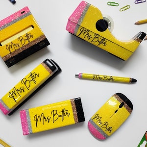 May include: A collection of yellow, glittery pencil-themed desk accessories, including a tape dispenser, pen, stapler, sticky note holder, and mouse. Each item is decorated with pink, silver, and black accents and the name "Mrs. Butter".
