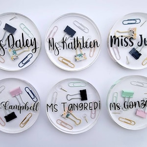 May include: Six clear resin coasters with embedded paper clips, binder clips, and push pins. Each coaster has a name in black script: Mrs. Daley, Ms. Kathleen, Miss Julia, Mrs. Campbell, Ms. Tangredi, and Ms. Gonzalez. The coasters are round.