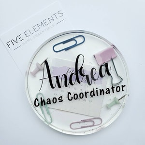 May include: A clear acrylic desk organizer with the name "Andrea Chaos Coordinator" written in black. The organizer has a calendar, push pins, paper clips, and a few other office supplies.
