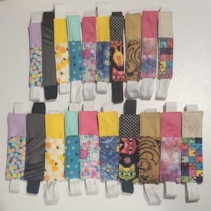 May include: A collection of 24 colorful fabric bookmarks with various patterns, including polka dots, flowers, cats, and geometric designs. The bookmarks are attached to white elastic bands.