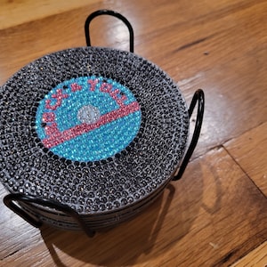 Puede incluir: Un juego de seis posavasos negros con un diseño de pedrería azul y rosa que dice "Gracias". Los posavasos están en un soporte de metal negro.